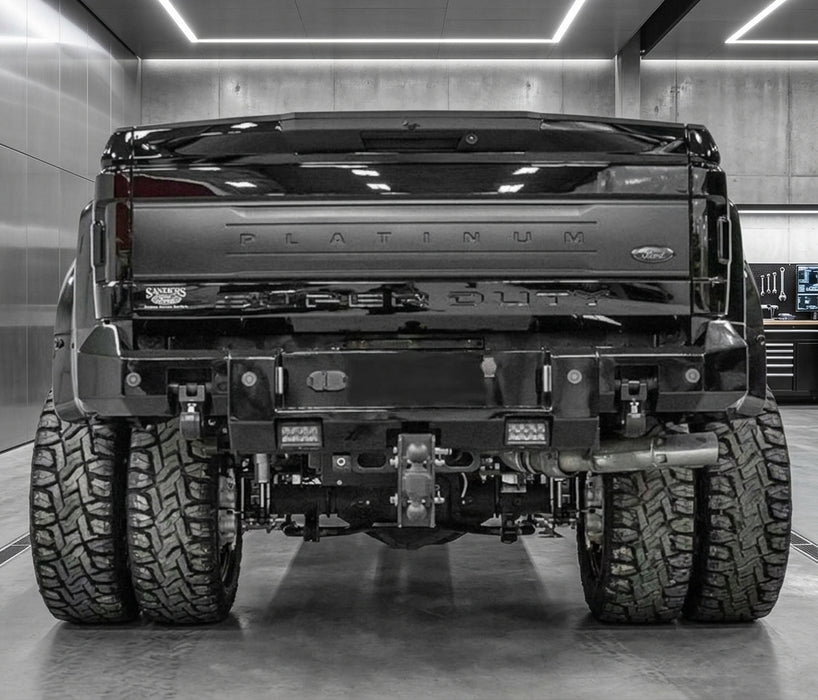 Rear view of a black Ford Platinum 3500 Super Duty truck with a heavy-duty custom rear bumper, dual rear tires, and off-road tires in a garage setting.